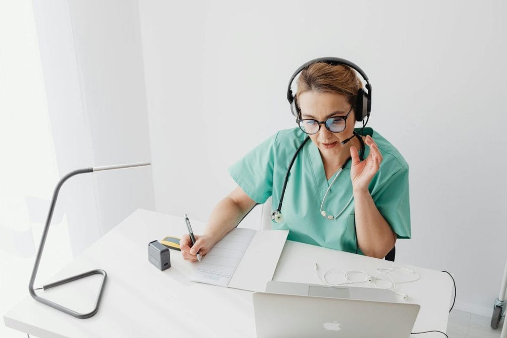 Doctor in scrubs with headset engages in an online consultation while taking notes.